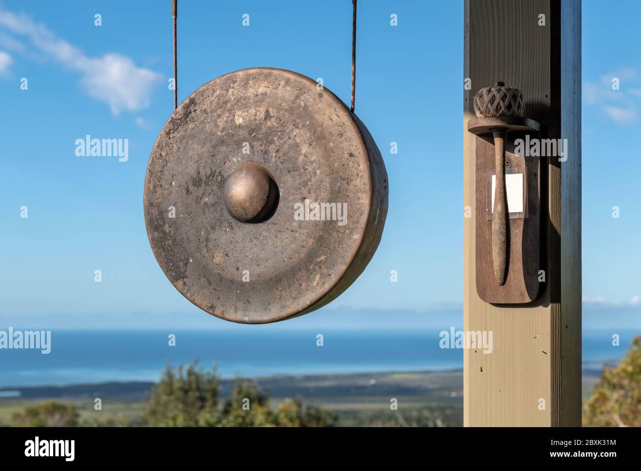 Un gong est suspendu à l'extérieur à côté d'un maillet monté sur un poteau.  L'arrière-plan est une vue sur l'océan, une végétation luxuriante et un  ciel bleu Photo Stock - Alamy