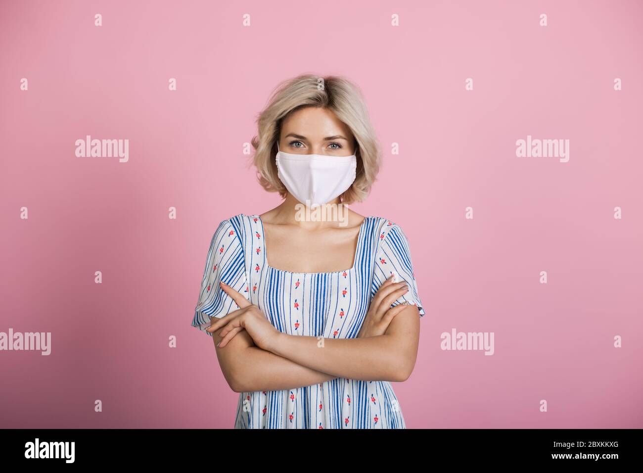 Blonde blanche femme portant un masque médical et une robe regarde la caméra sur un mur rose de studio Banque D'Images