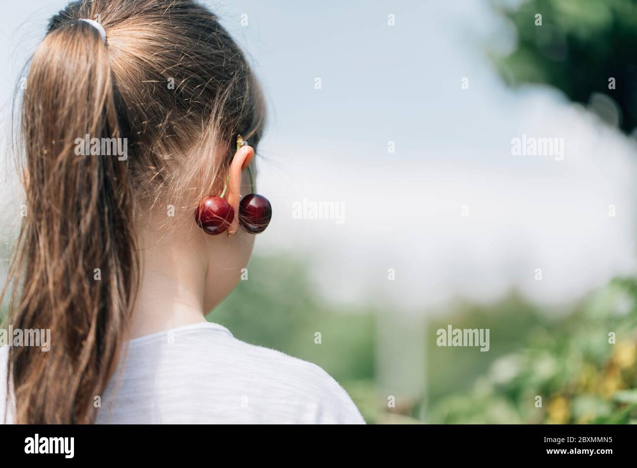 Belle petite fille a eu une cerise fraîche sur son oreille. Adorable petite fille avec une cerise sur l'oreille. Fabrication de bijoux à partir de fruits. Banque D'Images