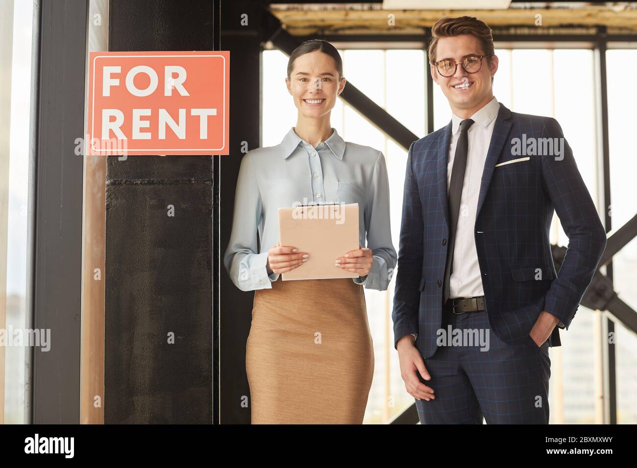 Portrait à la taille de deux agents immobiliers souriant à l'appareil photo tout en se tenant à côté de l'affiche ROUGE POUR LOUER, espace de copie Banque D'Images