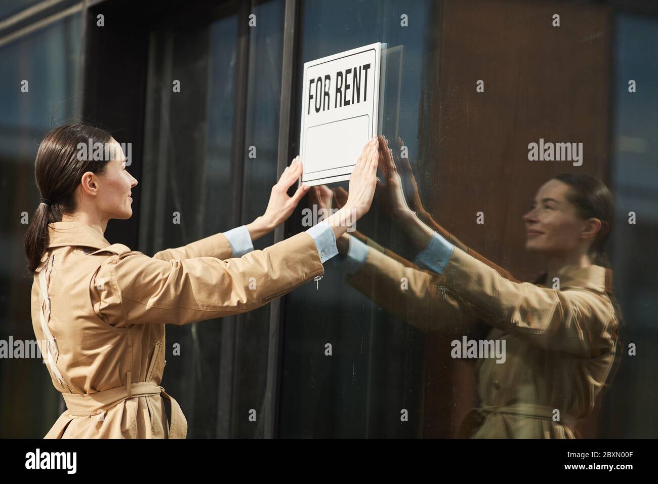 Portrait de la taille de l'agent de location de femme accroché POUR L'AFFICHE DE LOYER sur le bâtiment de bureau et souriant gaiement, espace de copie Banque D'Images