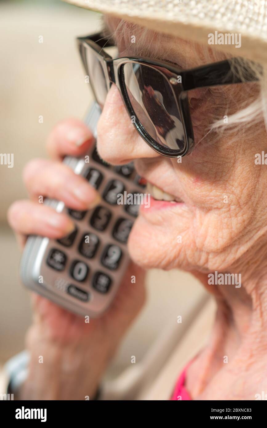 À son anniversaire, portant des lunettes de soleil et un chapeau d'été, assis dehors au soleil dans son jardin, en profitant d'un appel longue distance de sa petite fille. Banque D'Images