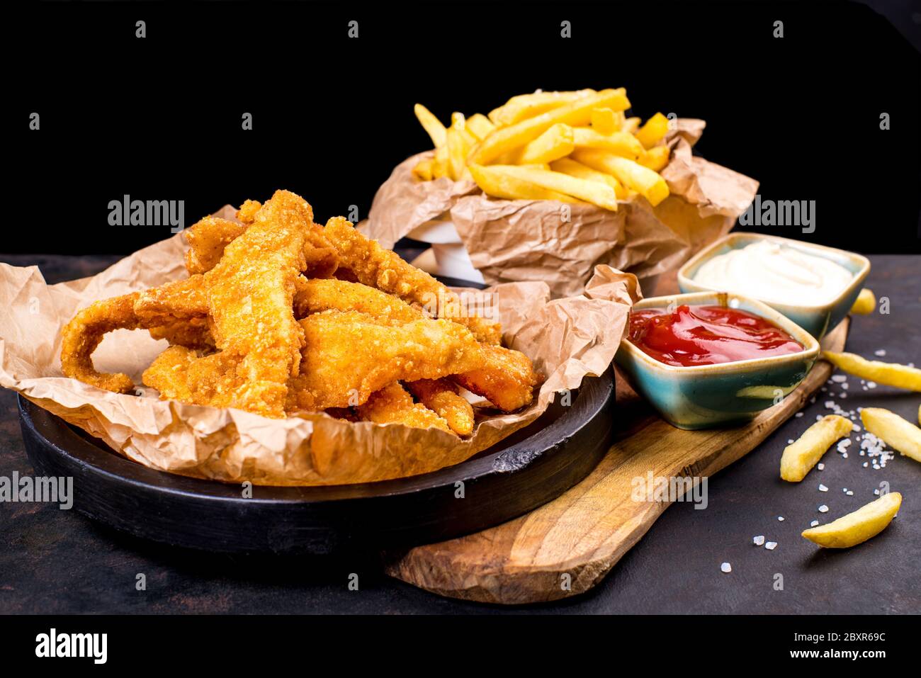 Beignets de poulet frits dans un seau noir avec sauce sur fond noir. Panés de poulet sur une assiette noire avec ketchup et frites. Banque D'Images