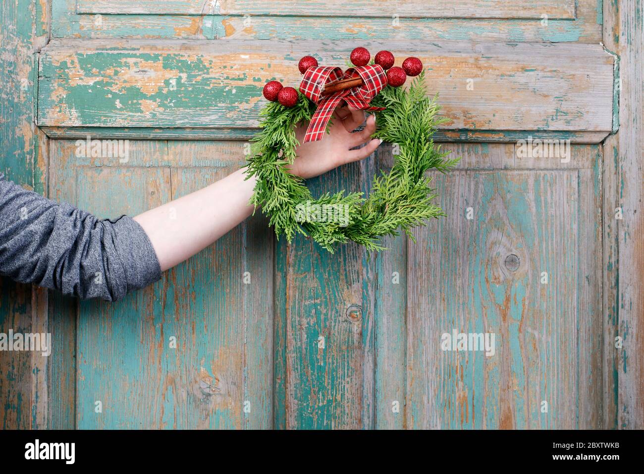 Femme tient une couronne de porte de noël traditionnelle avec des brindilles de thuja, des boules rouges et un ruban. Étape par étape, tutoriel. Banque D'Images