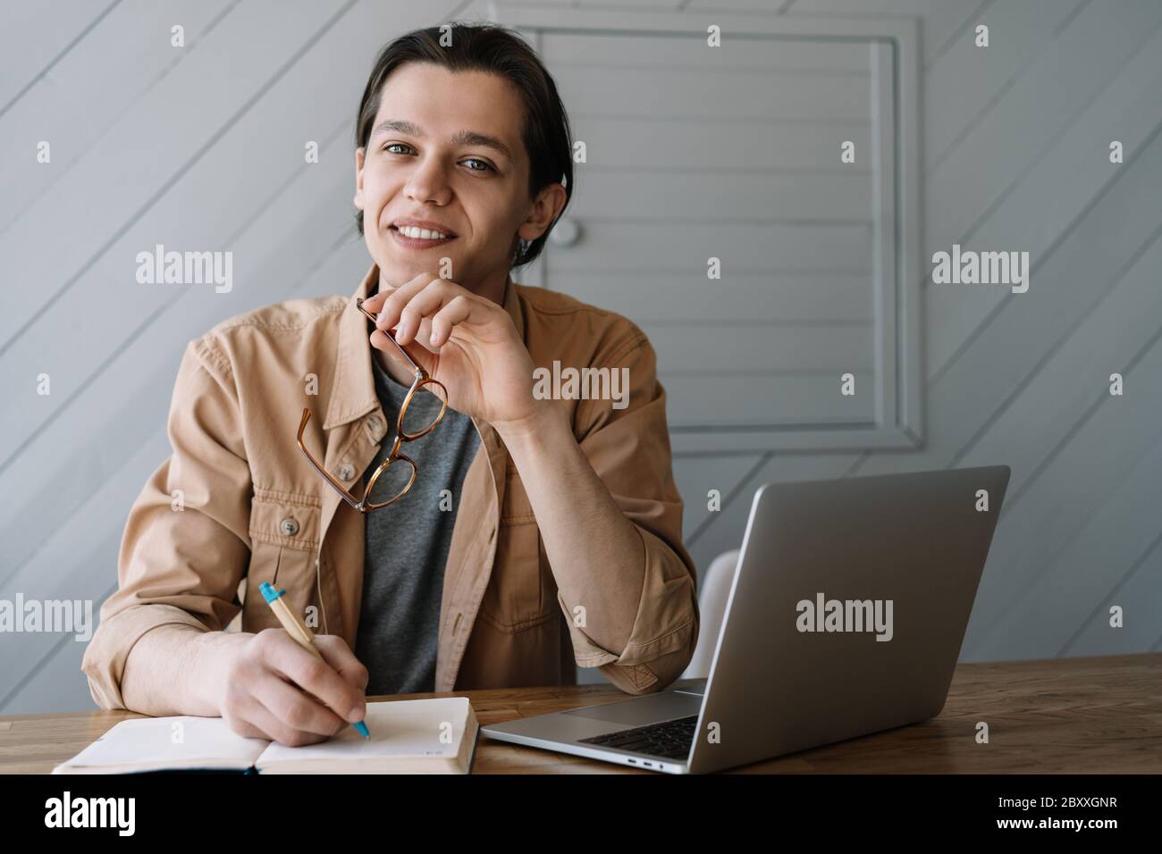 Un homme souriant écrit des notes dans un carnet, travaillant un projet indépendant dans un café. Jeune étudiant utilisant un ordinateur portable, étudiant, préparation à l'examen, apprentissage à distance Banque D'Images