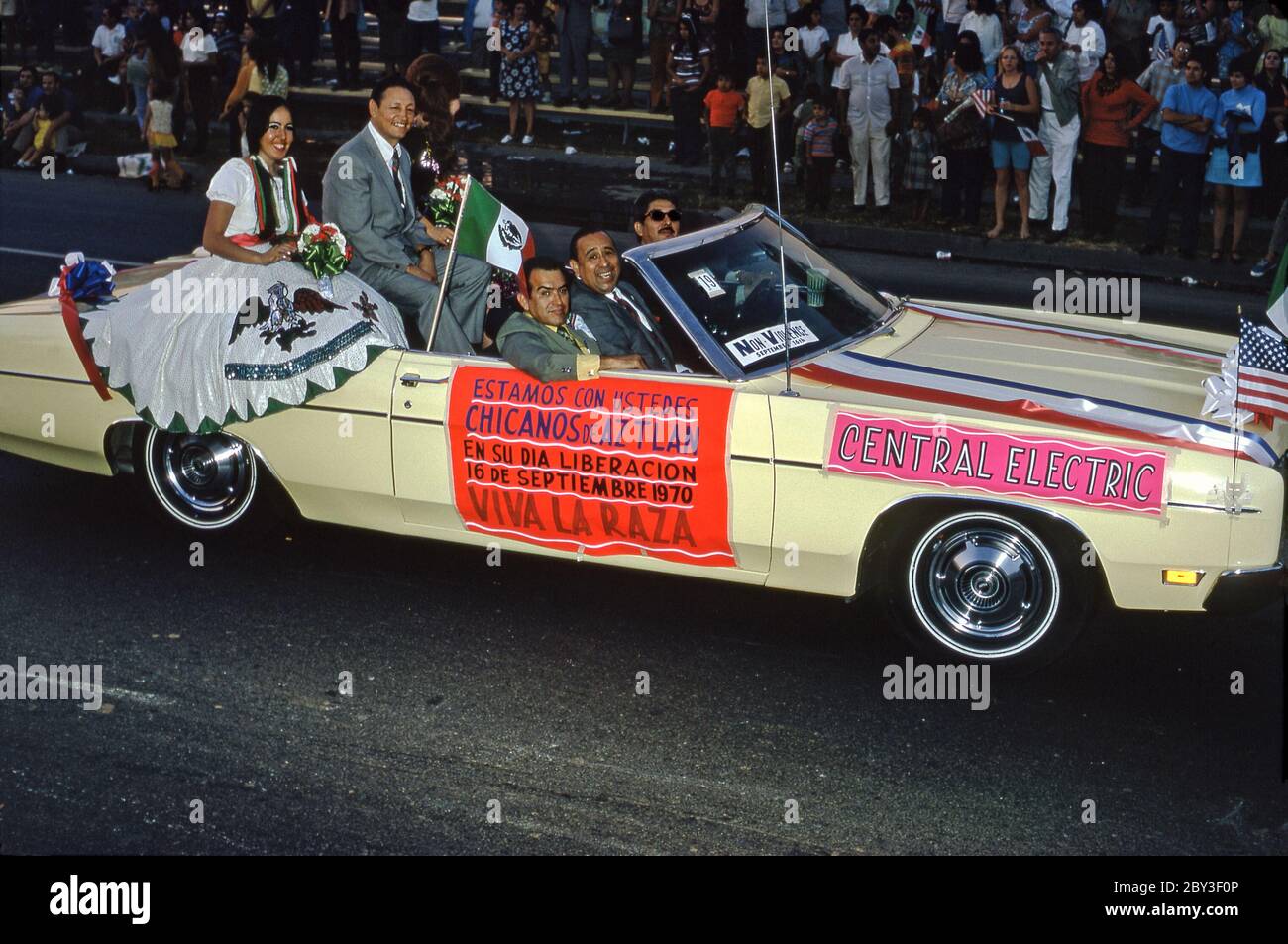 Une voiture dans un défilé de la communauté Chicano à Los Angeles, Californie, Etats-Unis 1970 (Chicanos et Chicanas sont des hommes et des femmes d'origine mexicaine). Une étiquette sur le côté de la voiture sponsorisée indique (en espagnol) la fierté de leur culture ('Viva la Raza' - vive la course). La Journée de l'indépendance (Día de la Independencia) est un jour férié au Mexique le 16 septembre pour célébrer la libération de la domination espagnole en 1810. Un drapeau mexicain vole aux côtés d'une femme en robe traditionnelle. Banque D'Images