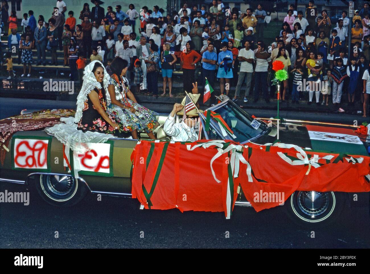Des couleurs vives sont utilisées pour décorer une voiture dans un défilé de la communauté Chicano à Los Angeles, Californie, USA 1970 (Chicanos et Chicanas sont des hommes et des femmes d'origine mexicaine). La Journée de l'indépendance (Día de la Independencia) est un jour férié au Mexique le 16 septembre pour célébrer la libération de la domination espagnole en 1810. Un drapeau mexicain vole aux côtés d'un drapeau américain et les couleurs vert blanc et rouge du drapeau mexicain dominent. Les femmes portent une robe traditionnelle aux couleurs vives. Banque D'Images