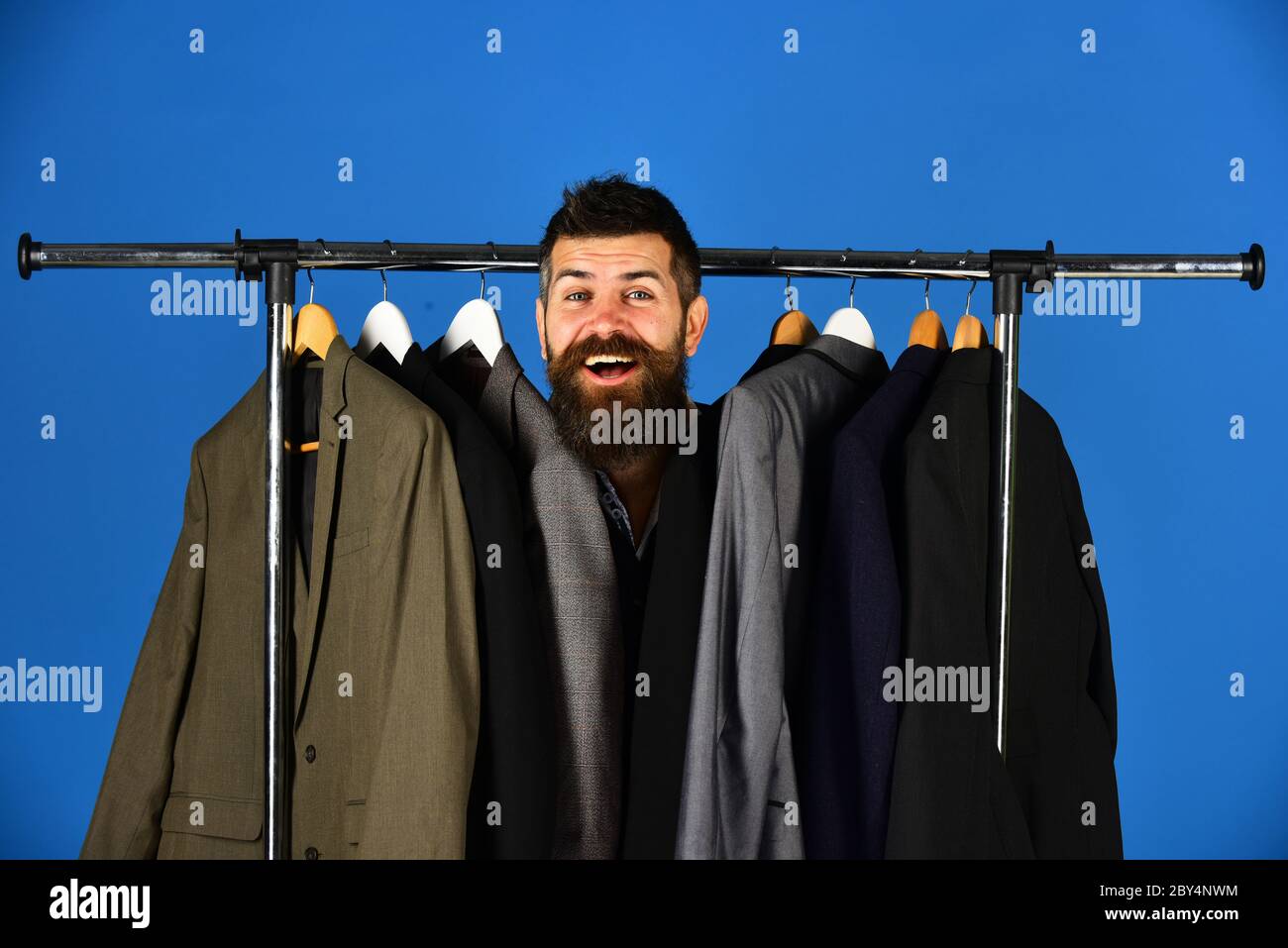 Portrait d un client masculin elegant par un porte vetements. Homme avec barbe et visage heureux regarde hors de l ensemble de vestes. Homme d affaires choisit un costume classique sur fond bleu. Sall...