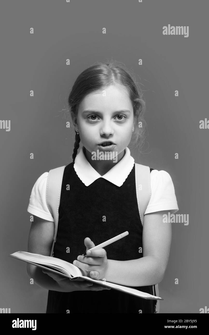 Enfant dans l'uniforme scolaire isolé sur fond bleu. Fille avec des tresses et une expression faciale attentive. La pupille contient un grand livre bleu et un marqueur vert. Étudier et de retour à l'école concept. Banque D'Images