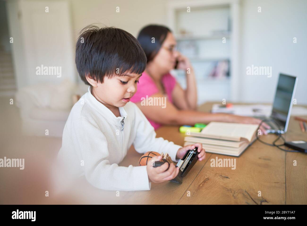 Garçon jouant avec l'agrafeuse à côté de la mère travaillant à l'ordinateur portable Banque D'Images