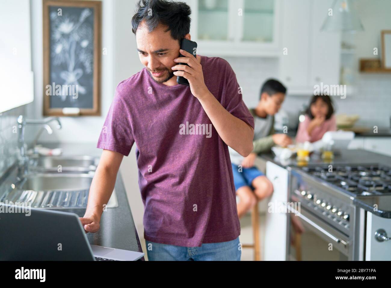 Homme travaillant à un ordinateur portable dans la cuisine avec des enfants Banque D'Images
