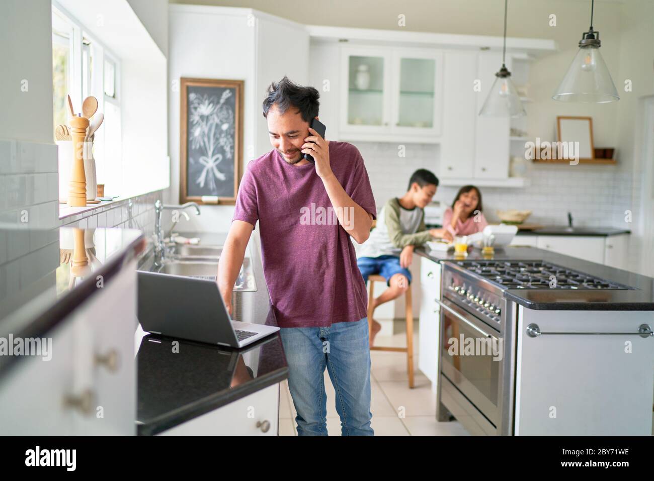 Homme travaillant à un ordinateur portable dans la cuisine avec des enfants qui mangent Banque D'Images