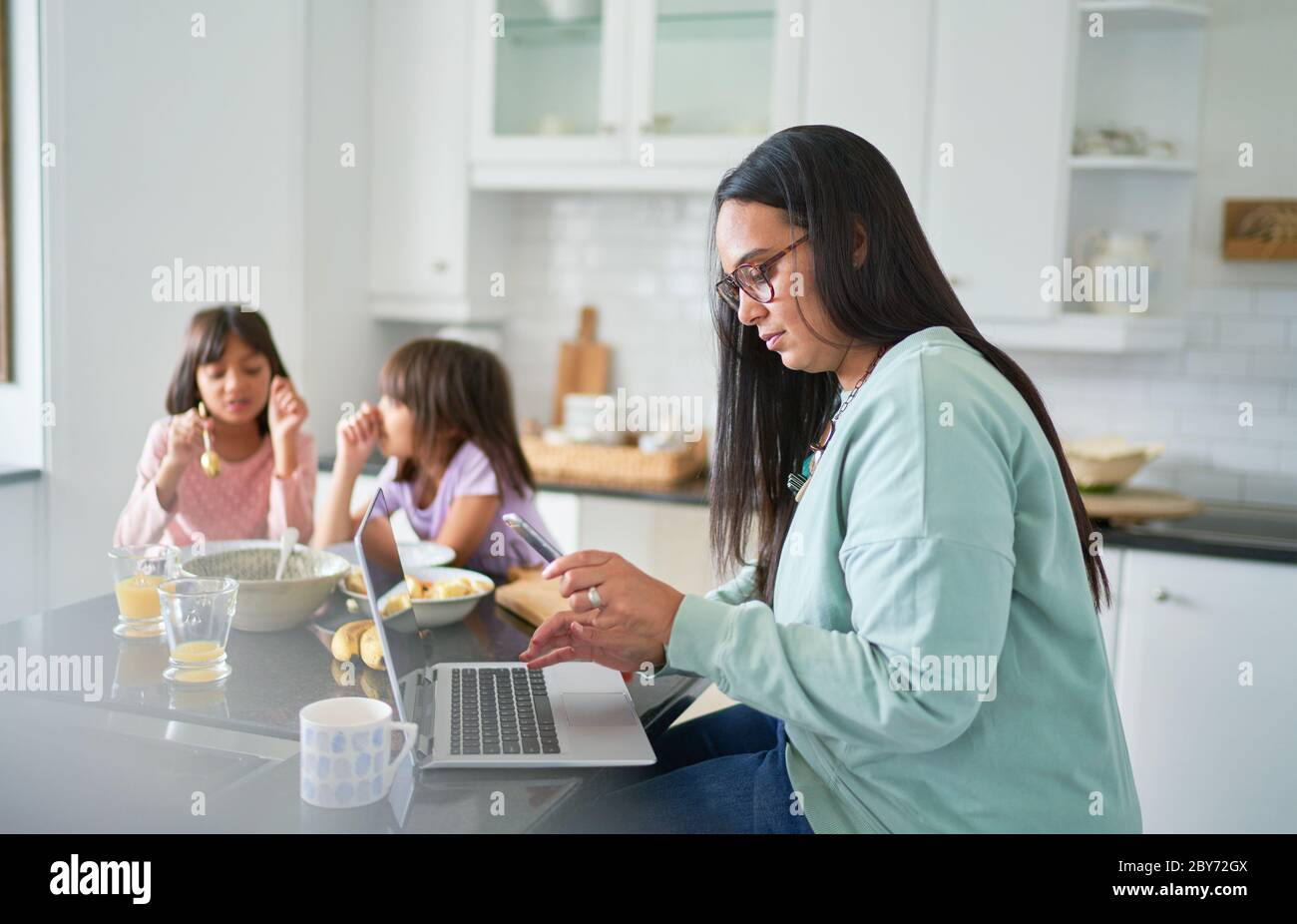 Mère travaillant à l'ordinateur portable dans la cuisine tandis que les filles mangent le petit déjeuner Banque D'Images