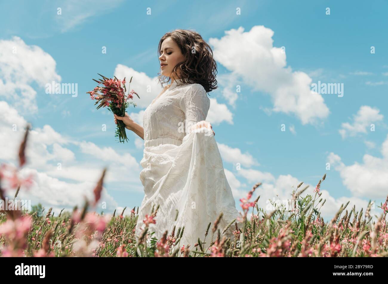 Portrait d'une jeune femme dans une longue robe blanche tenant un bouquet de  fleurs et sa robe comme elle marche dans un champ de fleurs contre le ciel  bleu Photo Stock -