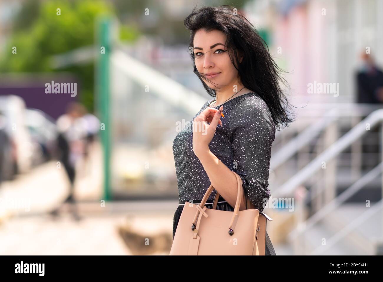 Élégante femme aux cheveux sombres qui marche à l'extérieur de la ville avec un sac à main en cuir sur son bras tournant pour regarder l'appareil photo avec un sourire amical et silencieux Banque D'Images