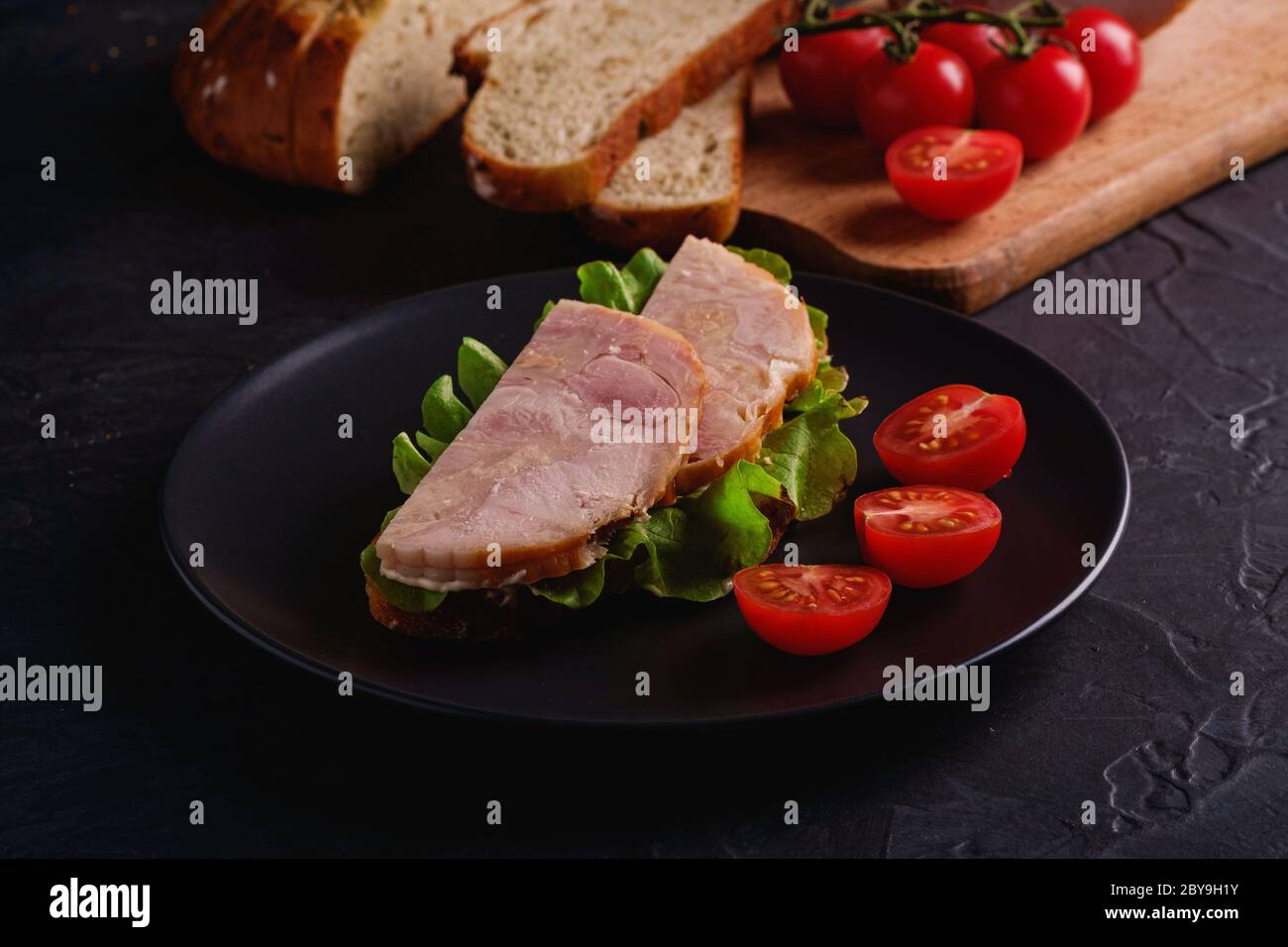 Sandwich avec de la viande de jambon de dinde, salade verte et des tranches de tomates cerises fraîches sur une assiette noire près des ingrédients sur la planche à découper Banque D'Images