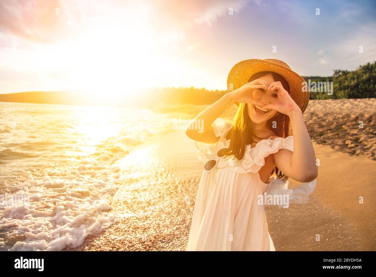 bonne jeune femme faisant un geste de coeur sur la plage au coucher du soleil Banque D'Images