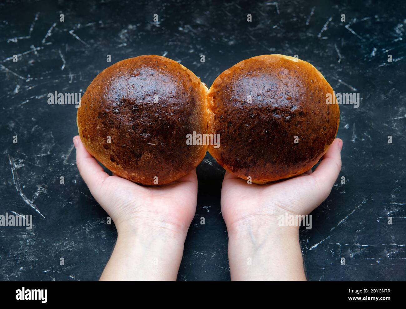Mains de femmes tenant deux petits pains dorés fraîchement cuits sur un fond noir texturé. Vue de dessus, mise au point sélective, prise de vue en studio. Plats maison et Banque D'Images