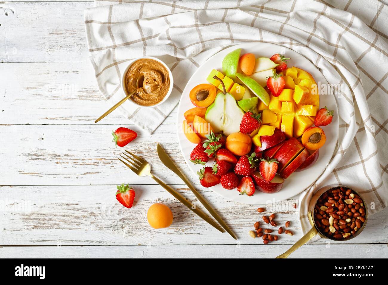 Dessert aux fruits colorés : plateau de fraises, mangue, pommes, poires, abricots, beurre d'arachide et arachides sur une assiette blanche sur une patte en bois blanc Banque D'Images