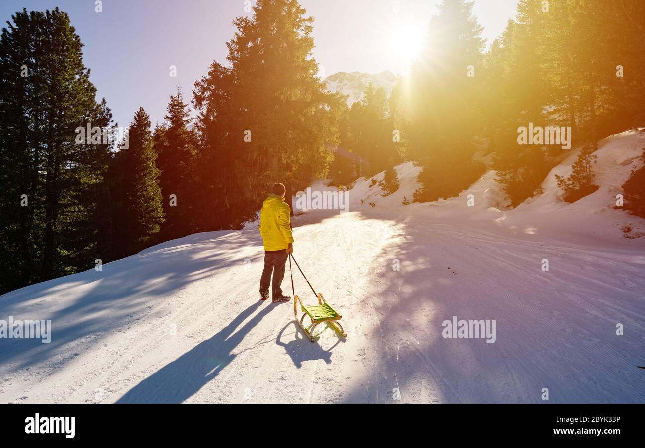 L'homme porte le traîneau sur la colline enneigée et profite du temps de luge d'hiver. Pris dans les Alpes au coucher du soleil Banque D'Images