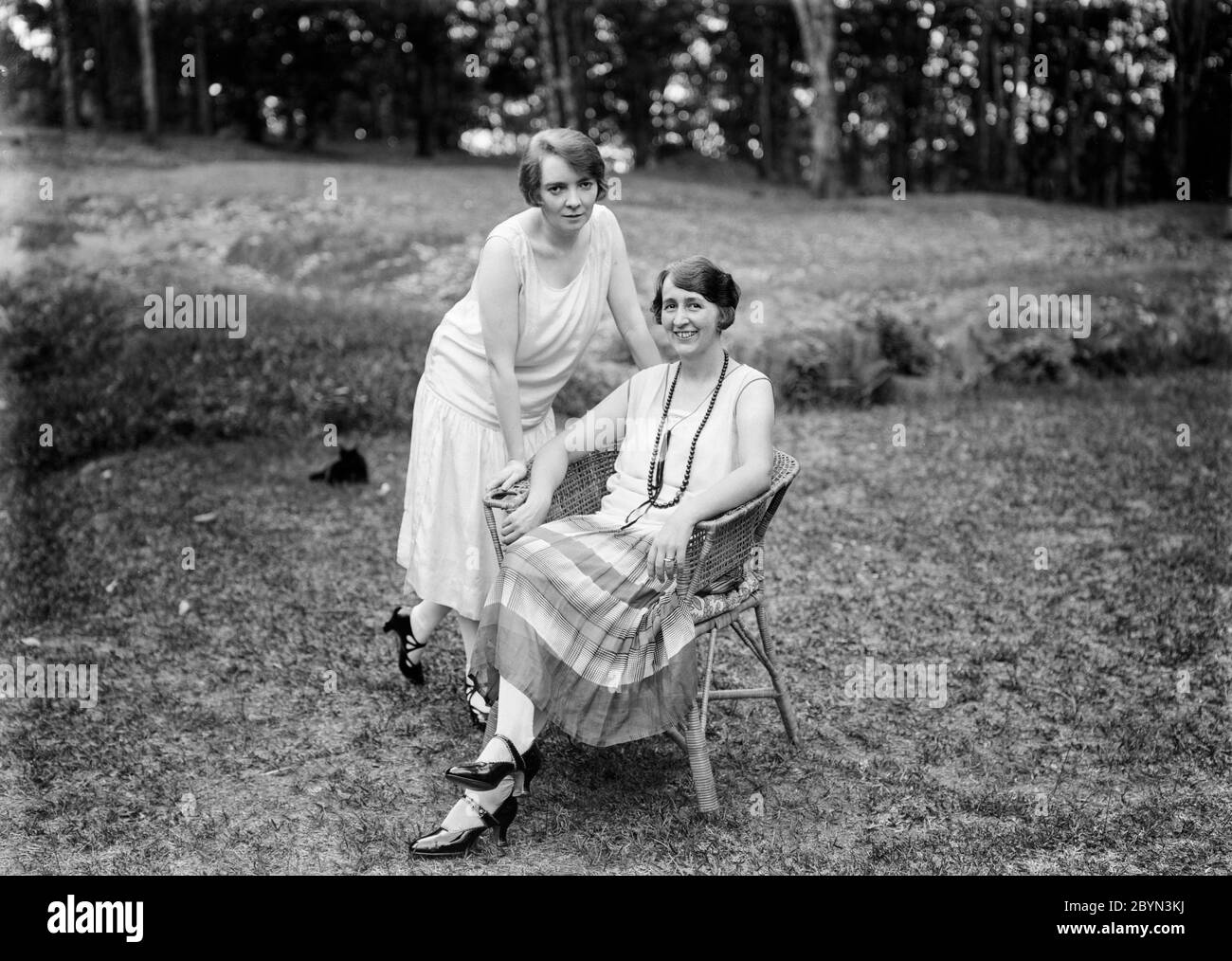 Photographie anglaise vintage des années 1920 en noir et blanc montrant une femme mûre, assise dans un jardin sur une chaise en osier, avec sa fille, une jeune femme, debout à côté d'elle, penchée sur la chaise. Les deux sont souriants et montrent la robe et la mode de l'époque. Banque D'Images