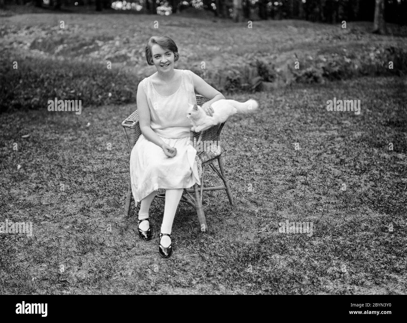 Photographie anglaise vintage noir et blanc du début du XXe siècle montrant une jeune femme, assise dans un fauteuil en osier, dans un jardin, tenant un chat. La femme montre la mode et le style de cheveux de l'époque, années 1920. Banque D'Images
