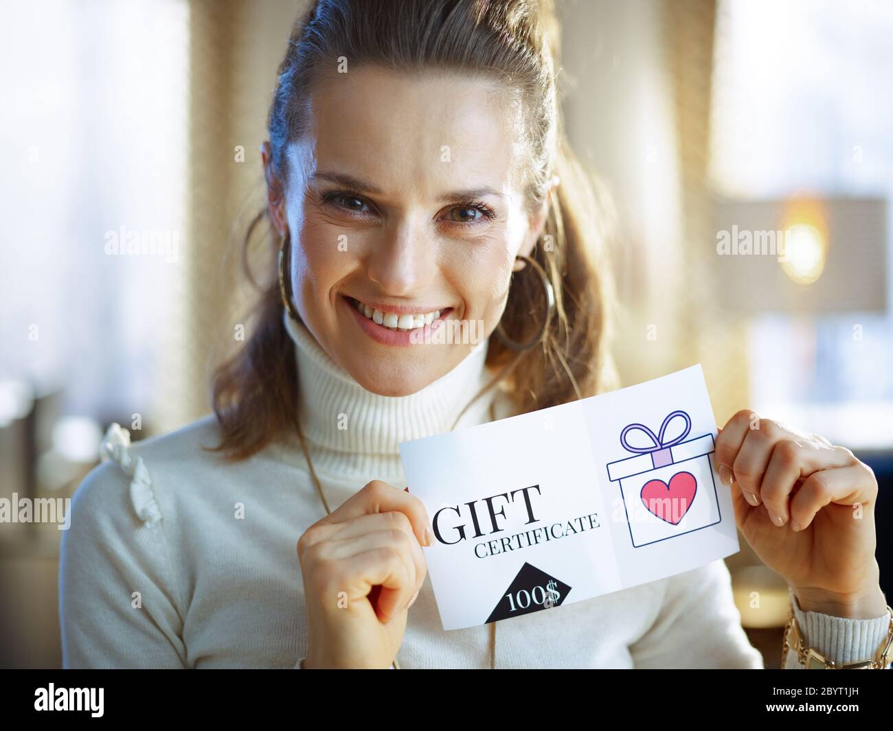 heureuse femme moderne en sweat blanc et jupe à la maison moderne en hiver ensoleillé avec certificat cadeau. Banque D'Images