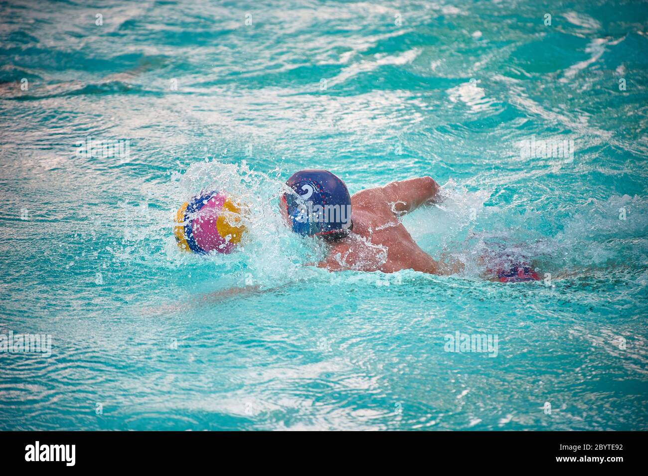 Un joueur de water-polo à la casquette bleue nageant dans une piscine après une balle éclaboussant de l'eau Banque D'Images