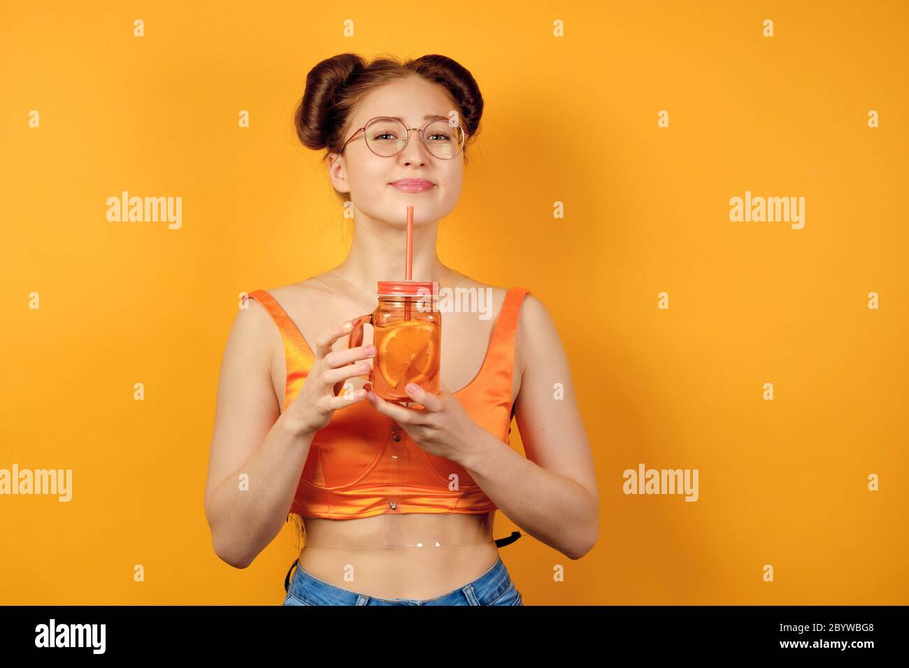 Une fille aux cheveux rouges en verres ronds et un haut orange est debout sur un fond jaune, souriant joli avec une limonade dans les mains Banque D'Images
