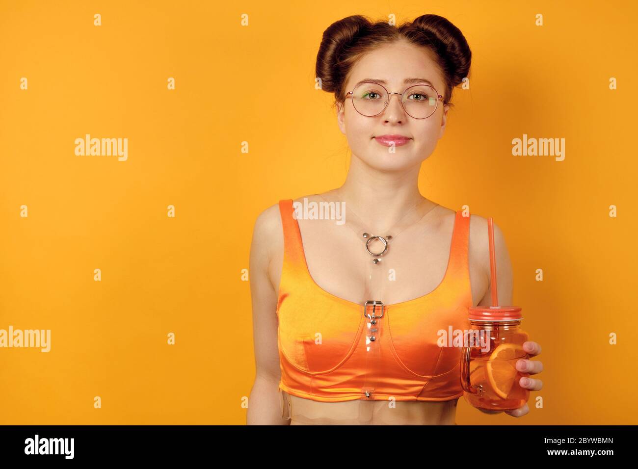 REDHEAD fille en verres ronds et un haut orange est debout sur un fond jaune avec un verre de limonade avec une paille dans les mains Banque D'Images