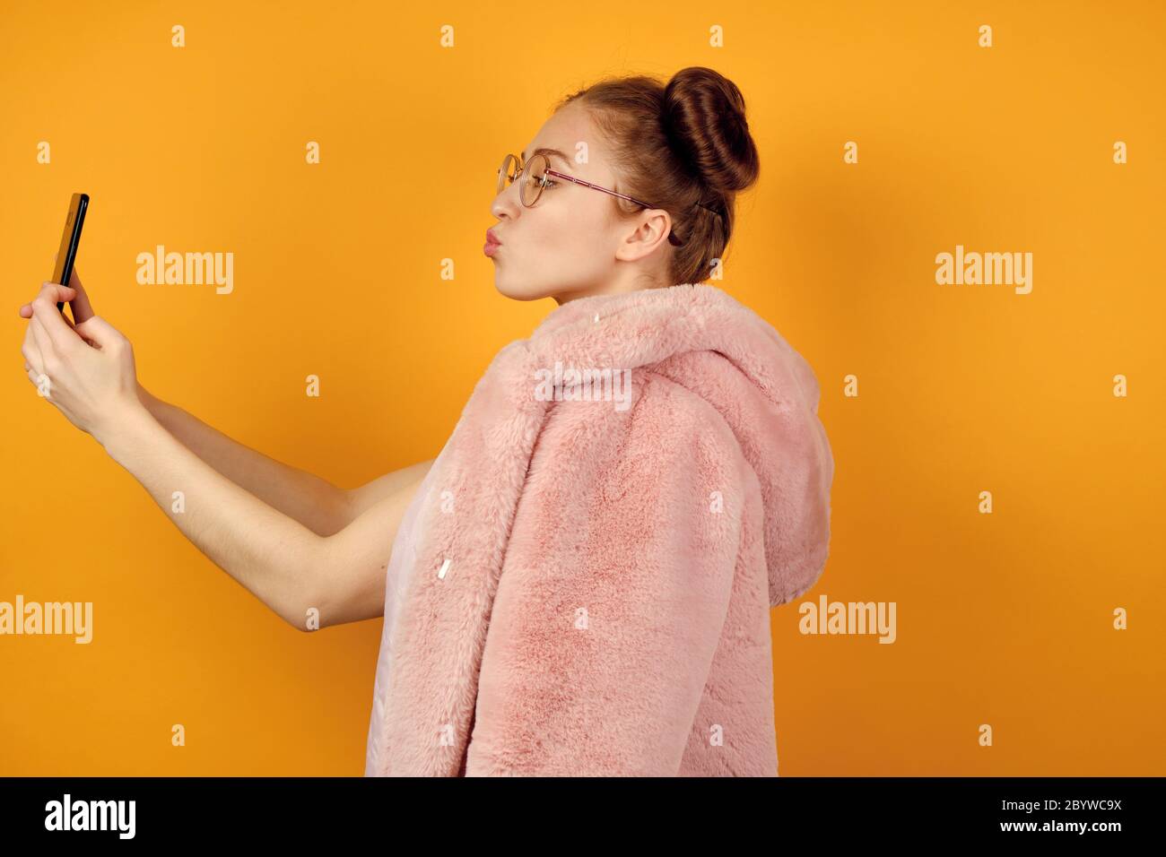 Une fille aux cheveux rouges en verres ronds se tient dans le profil dans un manteau rose et prend un selfie, étirant les lèvres dans un baiser Banque D'Images