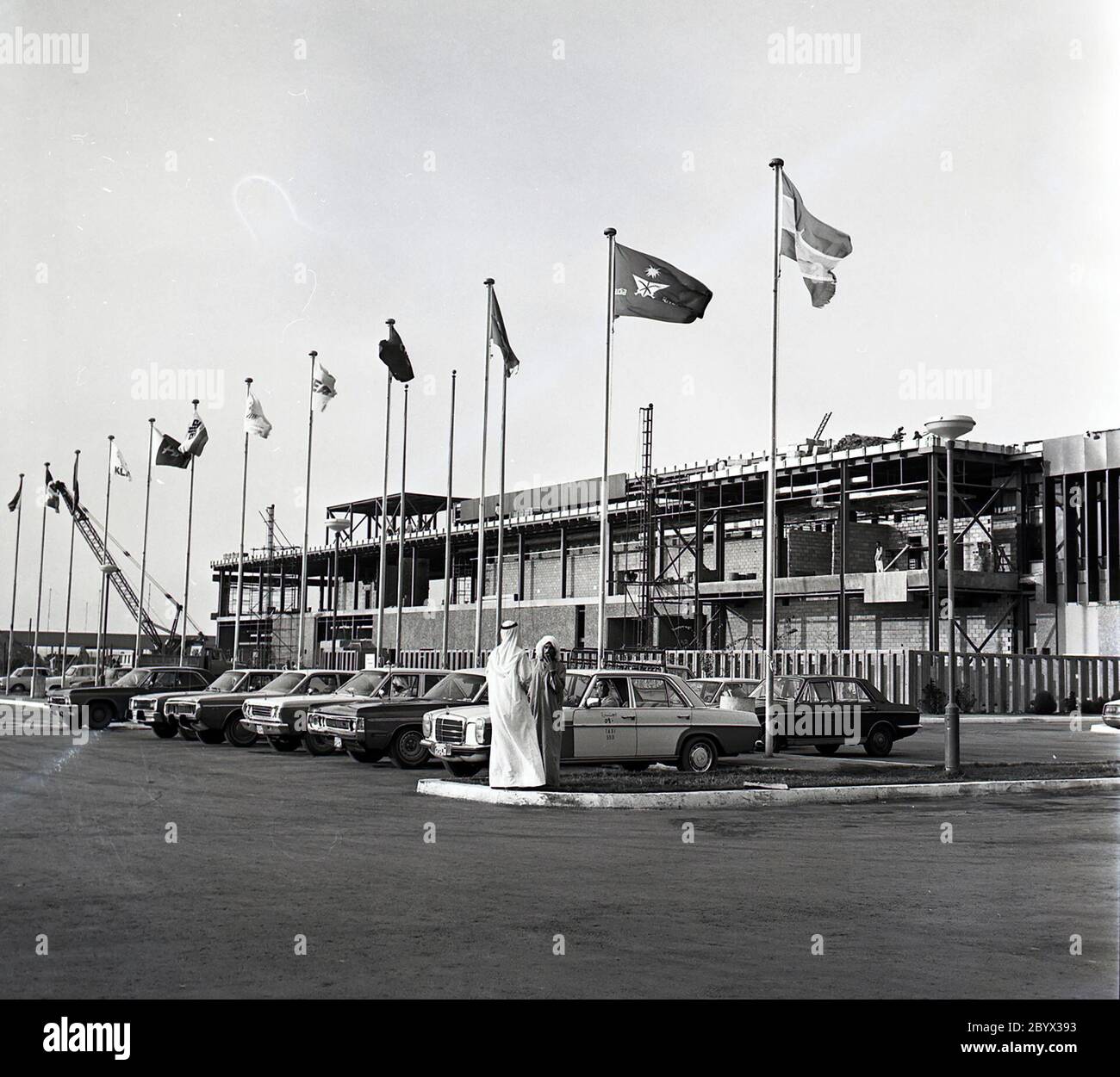 Une photographie de l'extérieur de l'aéroport international de Bahreïn en décembre 1975. Les taxis stationnés sont visibles au premier plan parmi les mâts de drapeaux Banque D'Images