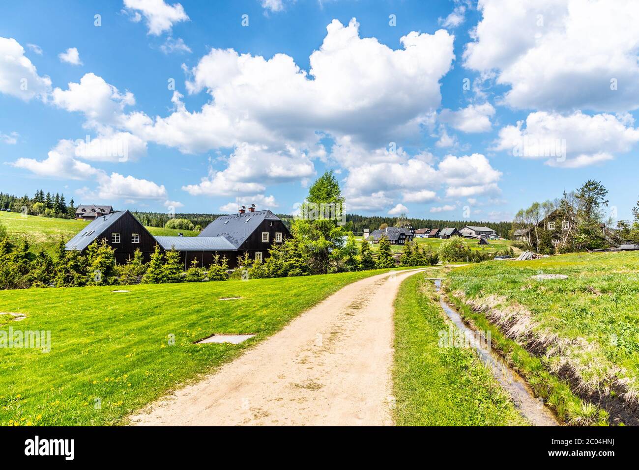 Journée d'été ensoleillée dans le village de montagne de Jizerka. Route de campagne poussiéreuse, prairies vertes et ciel bleu avec des nuages blancs, Jizera Mountains, République tchèque. Banque D'Images