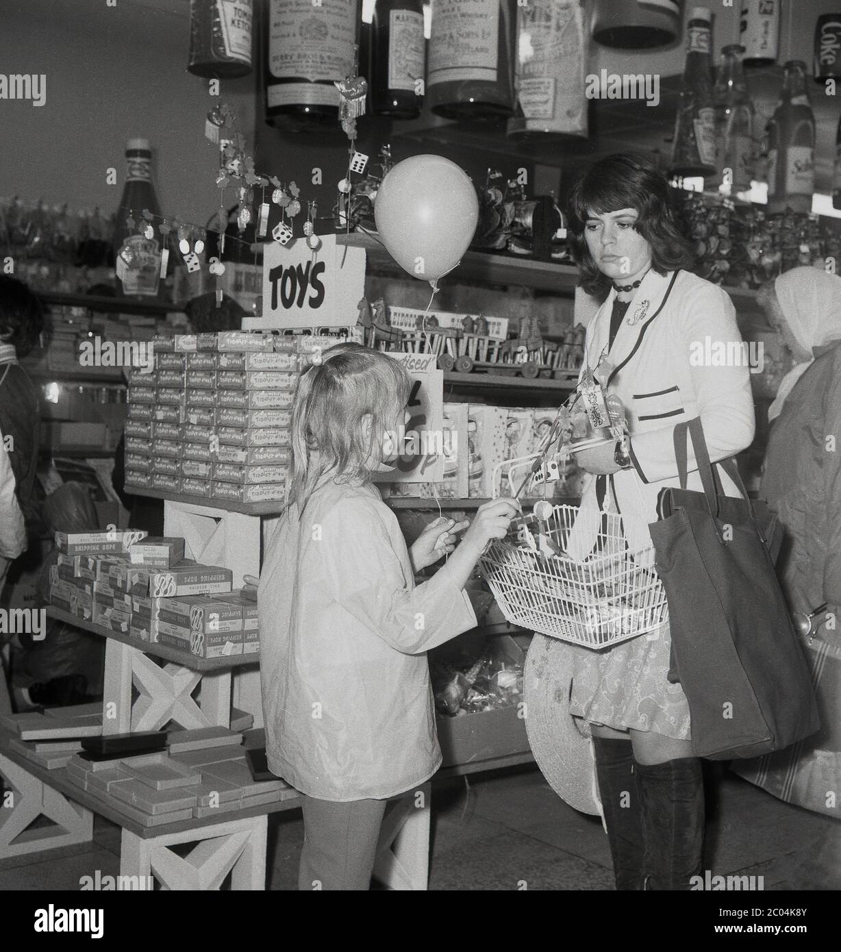 Années 1970, historique, une jeune mère avec un sac en toile et portant un panier de shopping en fil métallique avec sa fille dans un magasin de vente libre-service de l'époque vendant des jouets bon marché, des nicknacks et des produits de fantaisie, sud de Londres, Angleterre, Royaume-Uni. Banque D'Images