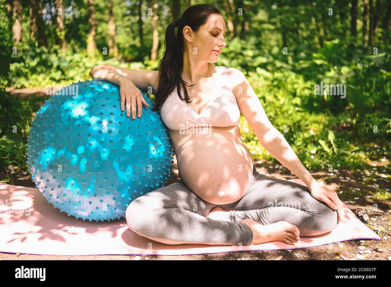 Belle jeune femme enceinte faisant de l'exercice avec la forme physique pilates Blue ball dans le parc extérieur. Asseyez-vous et détendez-vous sur un tapis de yoga rose. Un avenir actif Banque D'Images