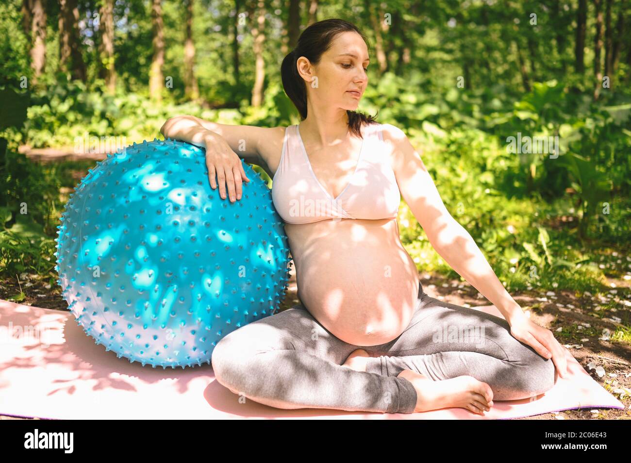 Belle jeune femme enceinte faisant de l'exercice avec la forme physique pilates Blue ball dans le parc extérieur. Asseyez-vous et détendez-vous sur un tapis de yoga rose. Un avenir actif Banque D'Images