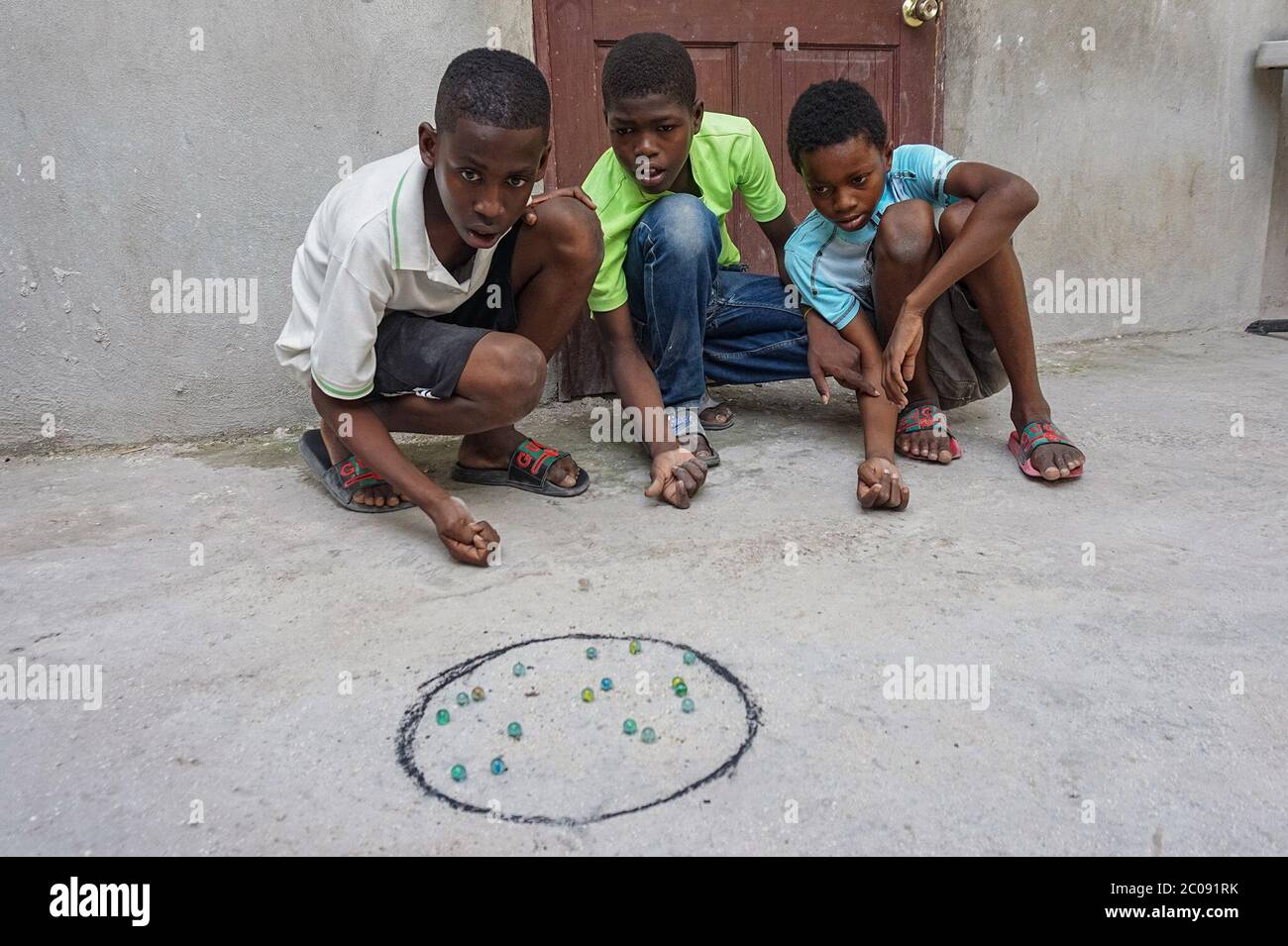 Guerby André, 14 ans (de gauche à droite), se concentre sur sa prochaine étape dans un jeu de marbres avec ses amis scolaires Gardy Mezil, 11 ans, et Cherdnerson Jean, 13 ans, à Port-au-Prince, Haïti. Le jeu est populaire chez les garçons ici, qui jouent souvent pendant les vacances scolaires. (Anne Myriam Bolivar, GPJ Haïti) Banque D'Images