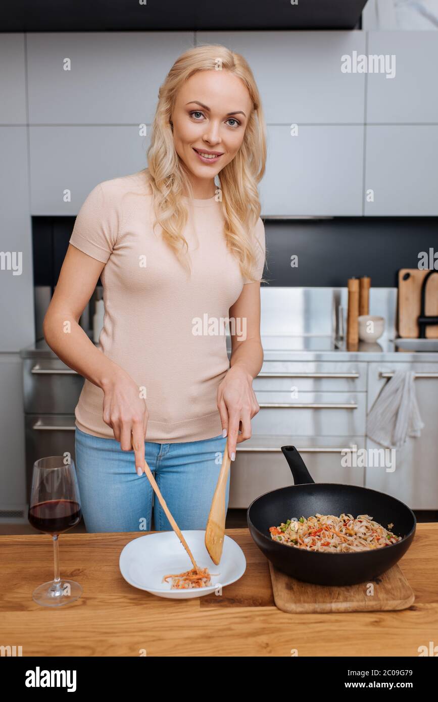 femme souriante mettant des nouilles thaïlandaises sur une assiette avec des spatules en bois tout en regardant l'appareil photo Banque D'Images