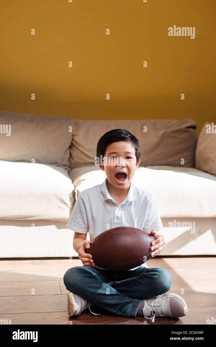 un garçon asiatique enthousiaste avec un ballon de rugby criant et regardant un match sportif à la maison en quarantaine Banque D'Images