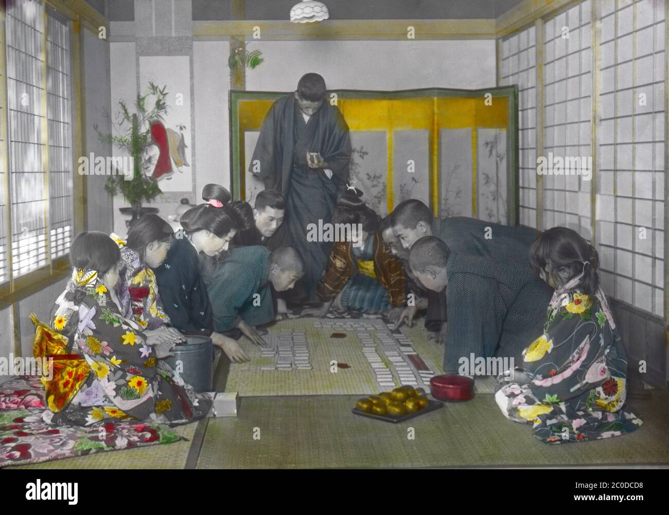 [ 1900s Japon - jouer à Karuta au nouvel an ] — membres de la famille jouant à Karuta au nouvel an. Une personne lit une partie d'un poème sur une carte yomifuda (lecture), tandis que d'autres tentent d'être les premiers à obtenir les cartes torifuda correspondantes (saisie) avec le reste du poème. Les poèmes sont issus de l'anthologie japonaise classique d'une centaine de waka japonais par une centaine de poètes connus sous le nom d'Ogura Hyakunin Isshu (小倉百人一首). Le jeu est toujours joué pendant le nouvel an aujourd'hui. diapositive en verre vintage du xxe siècle. Banque D'Images