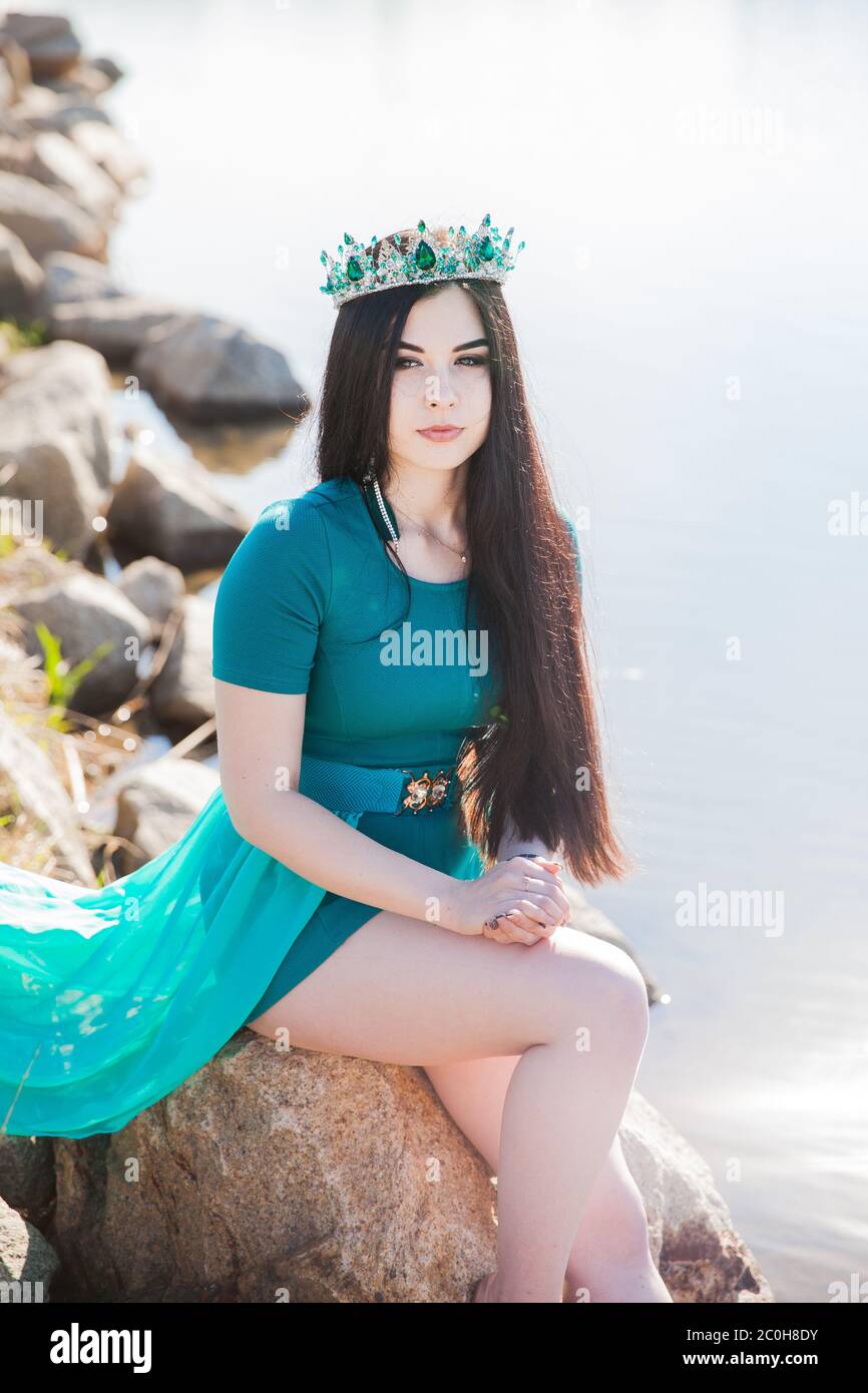 Queen dans une robe verte avec un train et un eorone émeraude. Brunette sexy sur le lac. Fille sur les pierres avec une grenouille. La nature féérique Banque D'Images