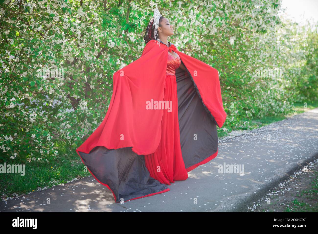 Reine dans la couronne et le manteau royal. Jolie fille sur le fond d'un jardin fleuri. Afro-américain en rouge Banque D'Images