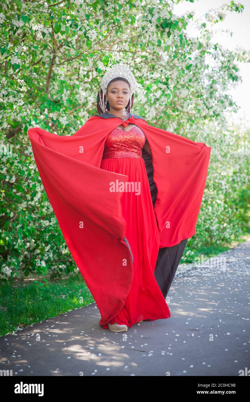 Reine dans la couronne et le manteau royal. Jolie fille sur le fond d'un jardin fleuri. Afro-américain en rouge Banque D'Images