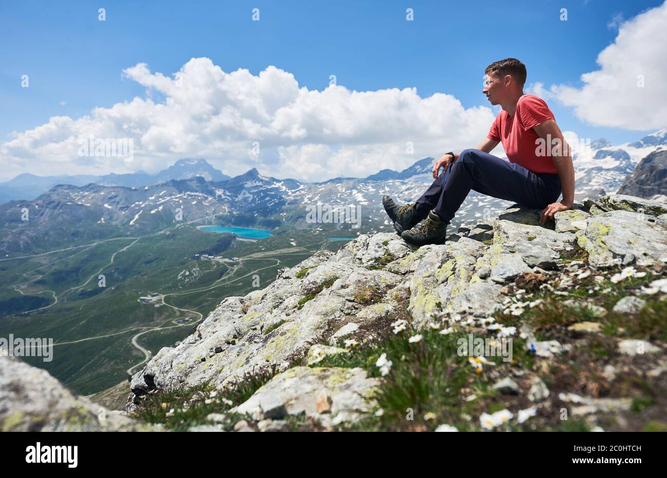 Vue latérale de touriste mâle assis sur le bord de la colline rocheuse sous ciel nuageux. Mountaineer en admirant la vue sur la vallée de montagne avec des collines herbeuses. Concept de voyage, de randonnée et de tourisme. Banque D'Images