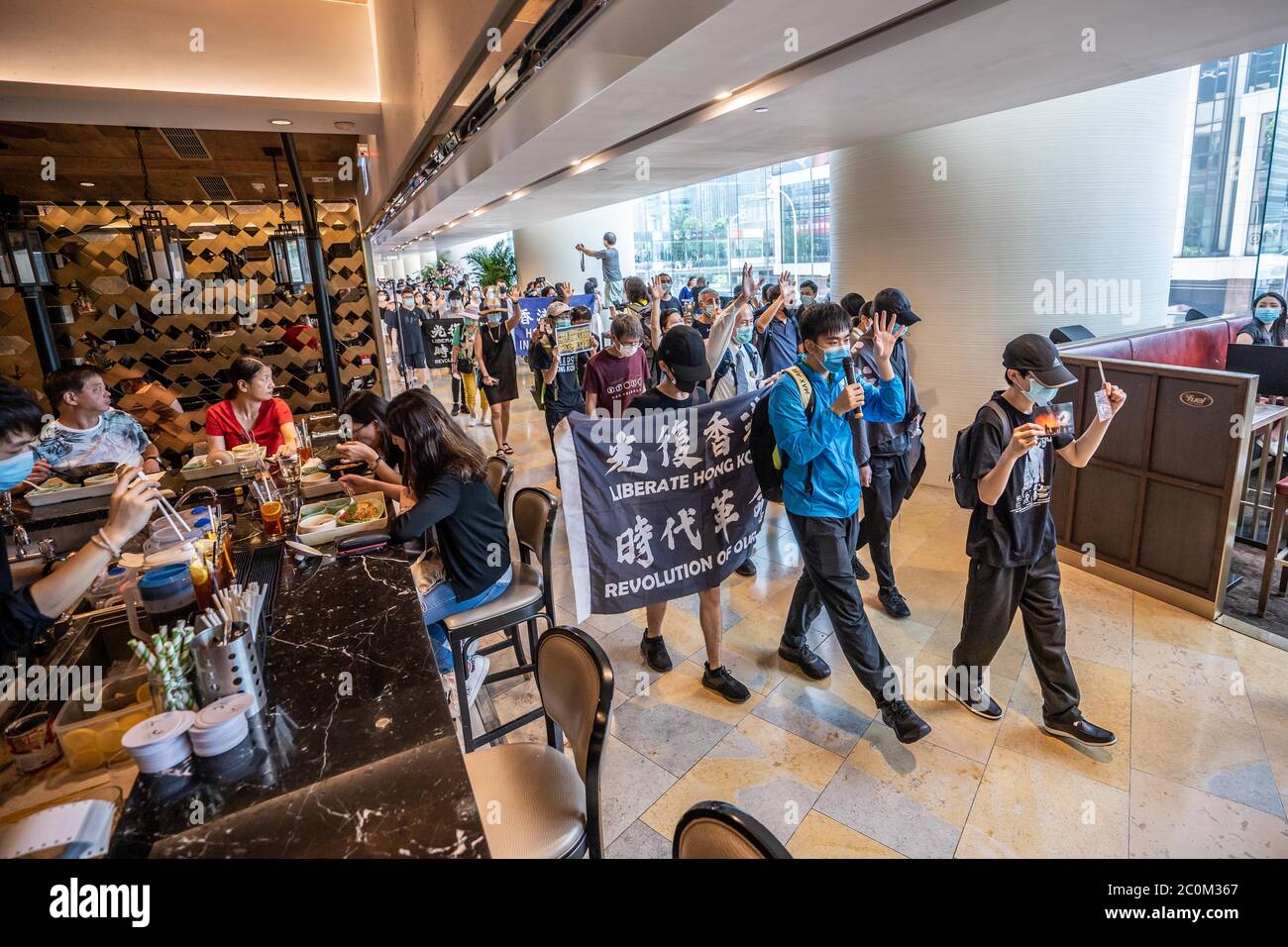 Les manifestants pro-démocratie défilent devant un restaurant tandis que les clients déjeunent à l'intérieur.les manifestants pro-démocratie ont organisé une manifestation dans le centre commercial Pacific place, dans le quartier central de Hong Kong, pour marquer le premier anniversaire du début du mouvement anti-gouvernement à grande échelle déclenché par le Le projet de loi d'extradition que beaucoup estiment sera utilisé comme un outil politique pour taire les ennemis politiques de la Chine à Hong Kong. Banque D'Images