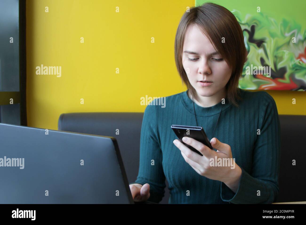Une fille au visage calme s'assoit à un ordinateur portable à une table dans un café, sur un canapé marron, contre un mur jaune. Elle tient dans sa main un smartphone, un téléphone mobile et regarde l'écran du gadget. Banque D'Images