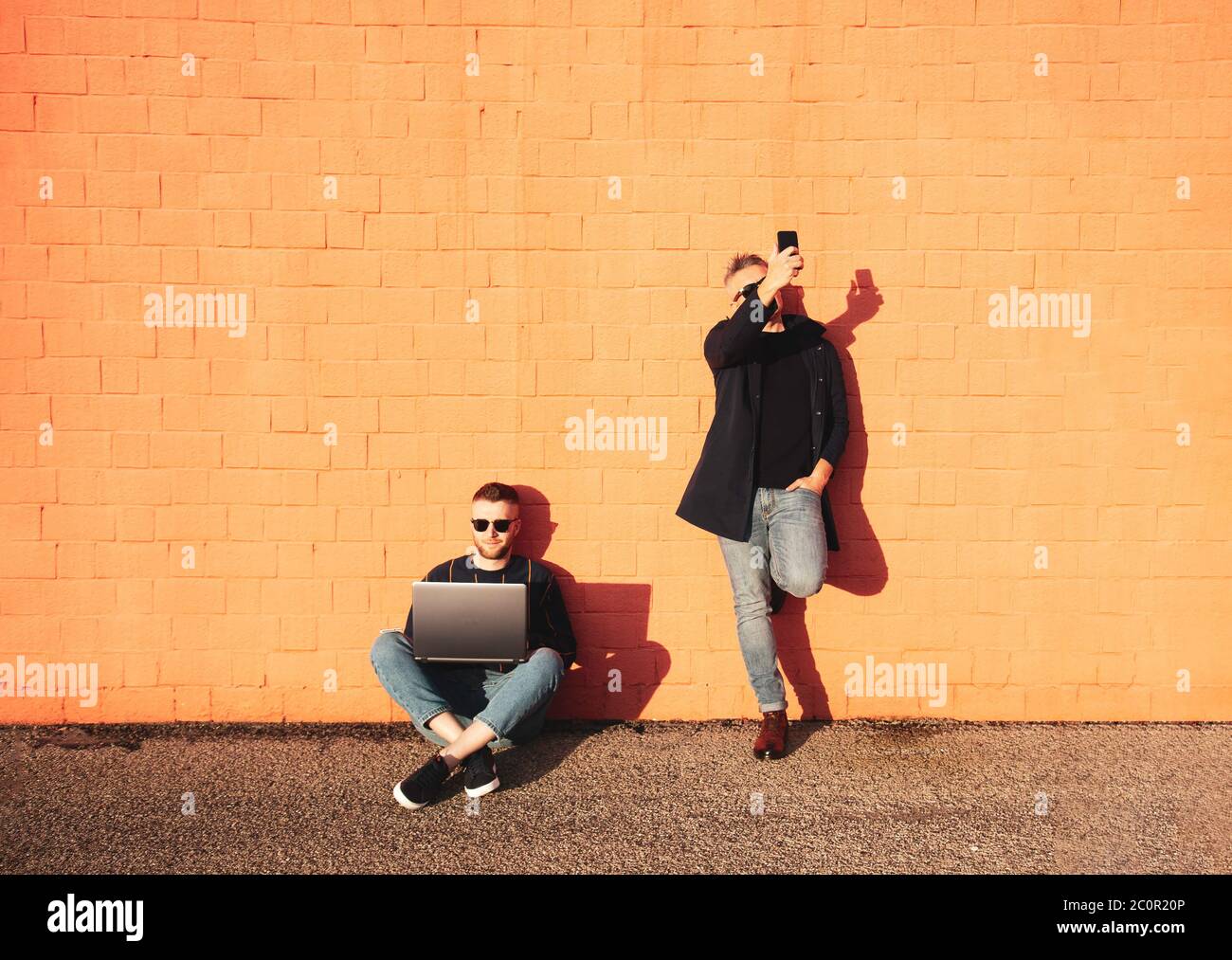 Deux amis ou collègues avec des appareils numériques pendant la pause déjeuner - jeune homme caucasien assis sur pieds croisés sur terre et travaillant sur ordinateur portable - l'autre Banque D'Images