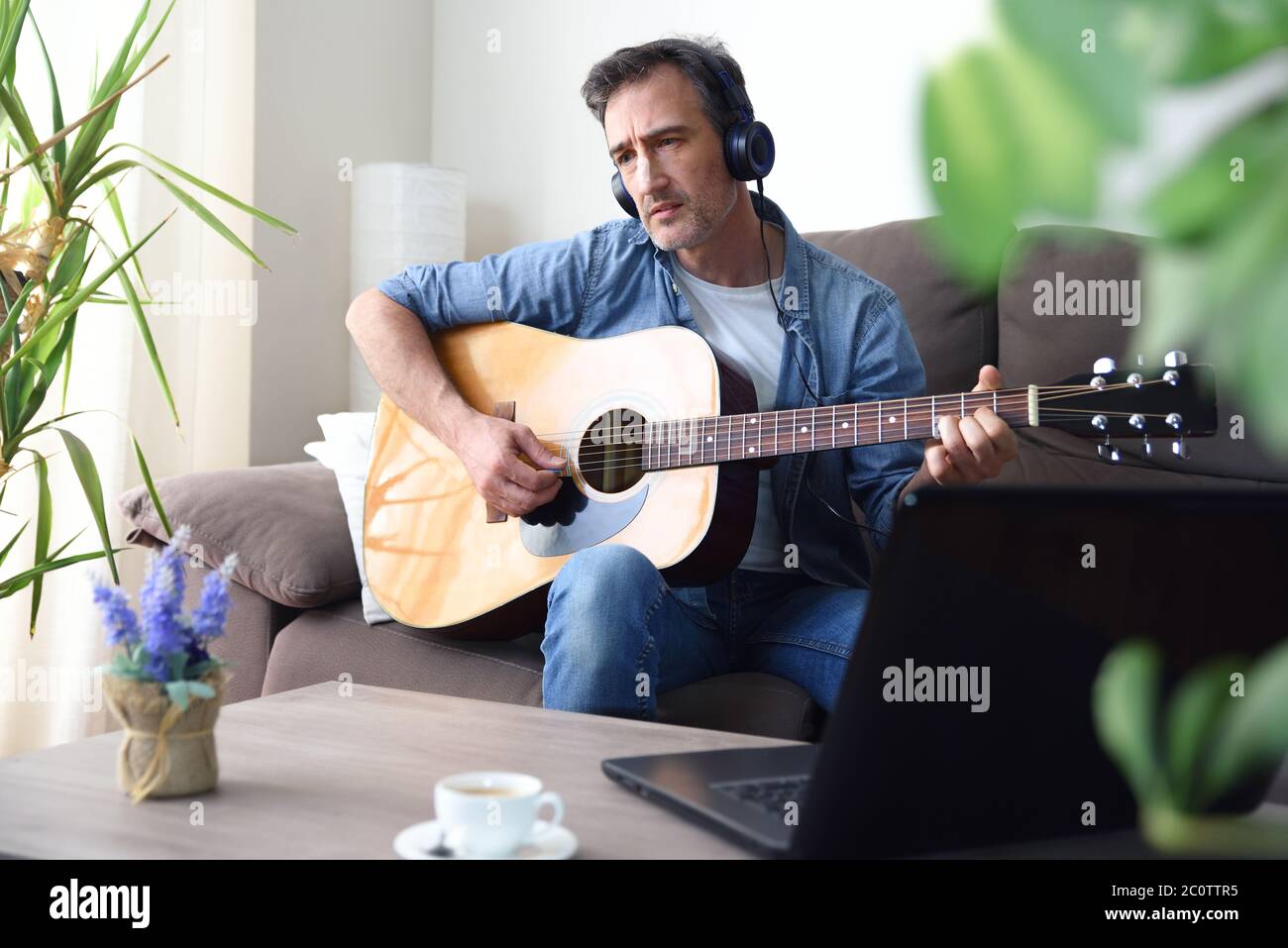 Homme écoutant de la musique depuis un ordinateur portable avec un casque et jouant de la guitare en même temps dans le salon à la maison Banque D'Images