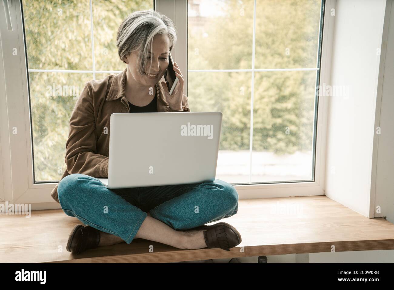 Une femme indépendante mature parle de téléphone portable, de travail ordinateur portable tout en étant assise sur mesure sur le fil de bois. Image teintée Banque D'Images