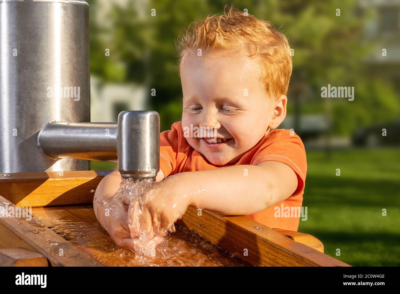 Un petit garçon souriant et heureux jouant avec de l'eau du robinet dans une aire de jeux ensoleillée Banque D'Images